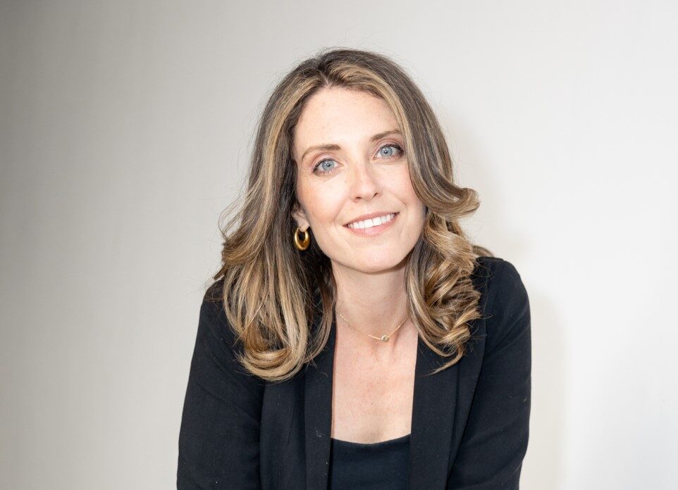 A woman in a black jacket and tan pants sitting on a white couch.